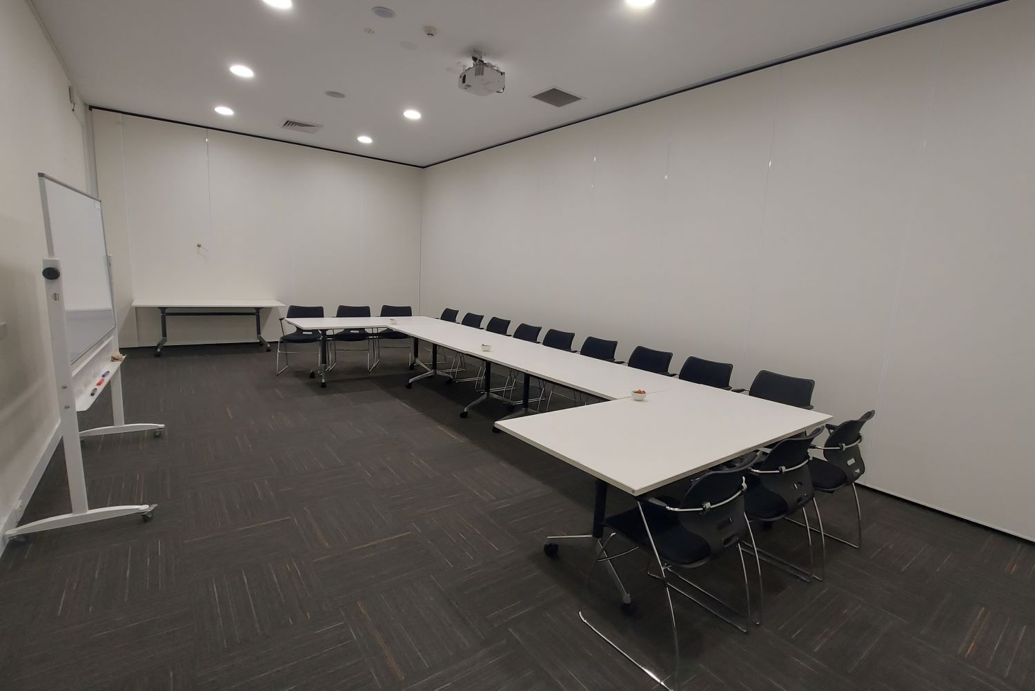 large meeting room with u-shape table layout, whiteboard, projector on roof. White walls, grey carpeted floor.