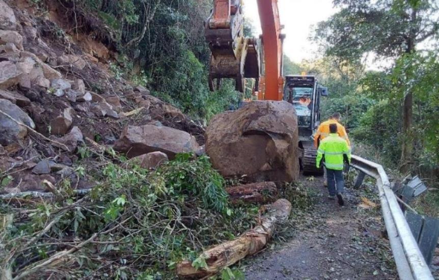 An excavator works to remove a large boulder and landslide debris covering a road.